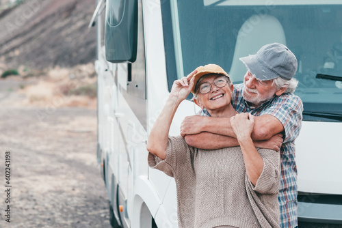 Happy senior couple on leisure trip enjoying travel destination standing outside a camper van motorhome hugging cuddling. Smiling elderly man and woman, transport and tourism concept lifestyle