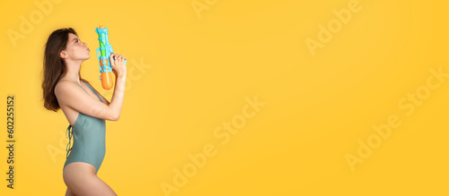 Cool young european woman in swim clothes holding water gun, having fun on yellow studio background, panorama copy space