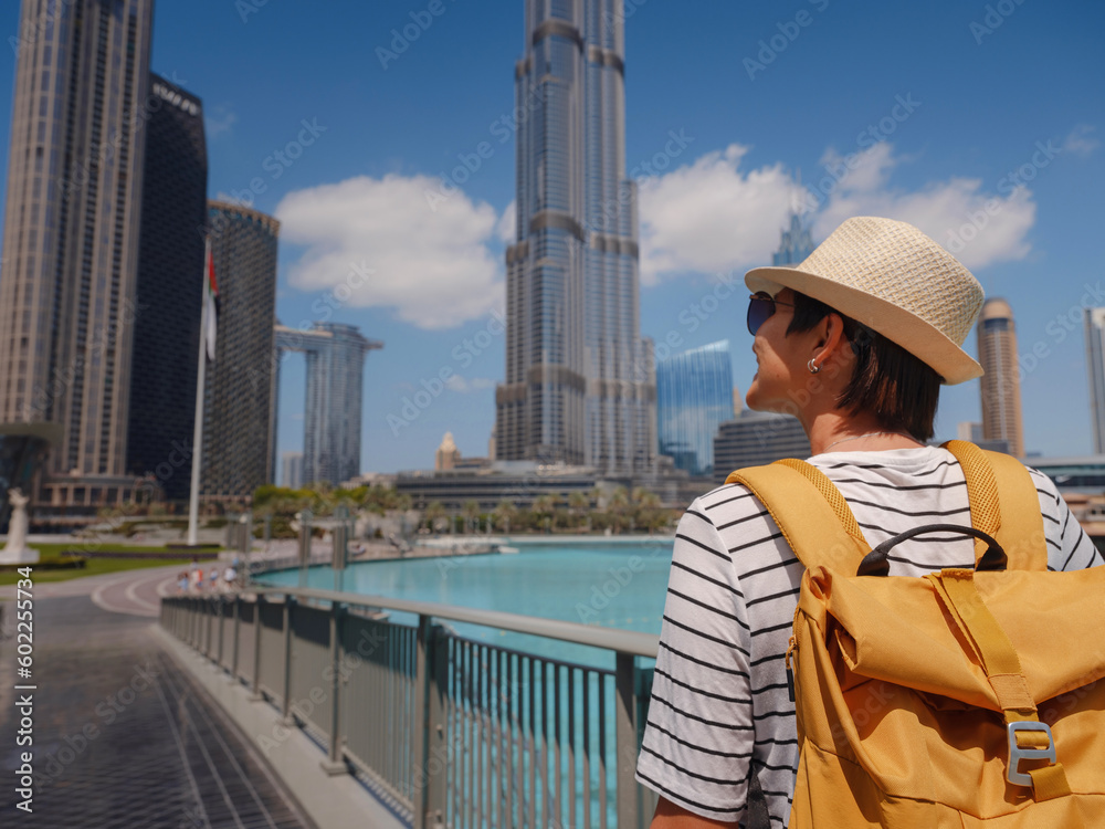Enjoying travel in United Arabian Emirates. Young woman with yellow backpack walking on Dubai Downtown in sunny summer day. view from the back or rear view