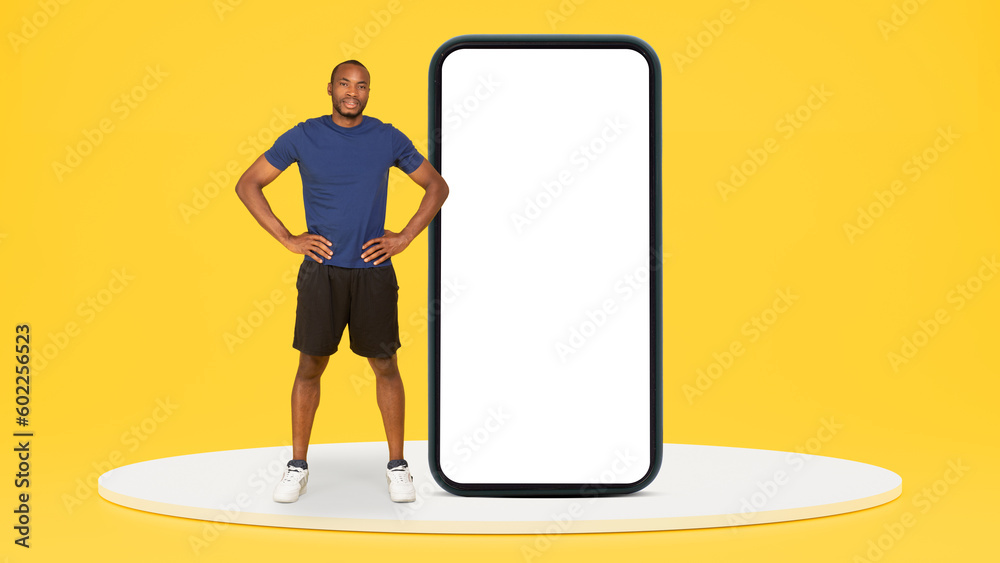 Smiling young black male athlete trainer in sportswear on platform near big smartphone with empty screen Smiling young black male athlete trainer in sportswear on platform near big smartphone with empty screen