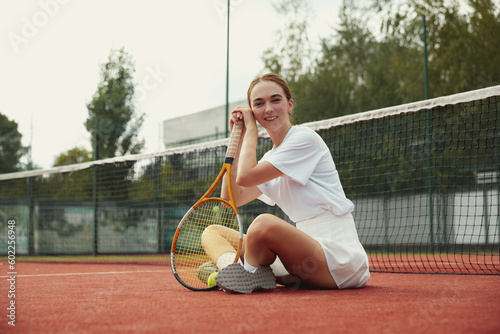 Young female tennis player in sportive white clothes sitting on the ground in the court at the tennis net at summer daytime. Grounded view. © Davidovici