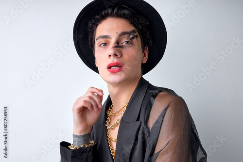 Sexy womanlike feminine male in black translucent shirt and hat posing at camera isolated over white studio background. handsome guy with chain and bright party make-up, looking passionate, sensual