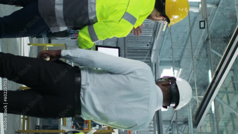 Vertical shoot. Full height engineers going at construction site. Workers are shooted from back and have discussion. Manufacture employee dressed up in reflective vest and hold tablet. Vertical shoot. Full height engineers going at construction site. Workers are shooted from back and have discussion. Manufacture employee dressed up in reflective vest and hold tablet.