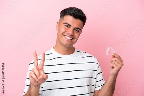 Young caucasian man holding invisaling isolated on pink background smiling and showing victory sign photo