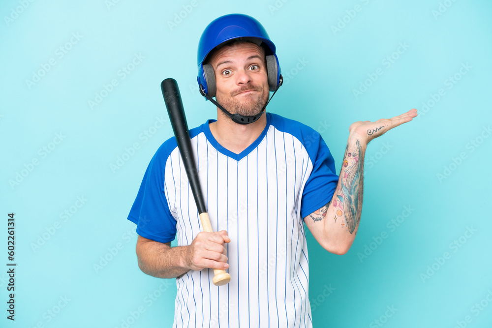 Baseball player with helmet and bat isolated on blue background having doubts while raising hands Baseball player with helmet and bat isolated on blue background having doubts while raising hands