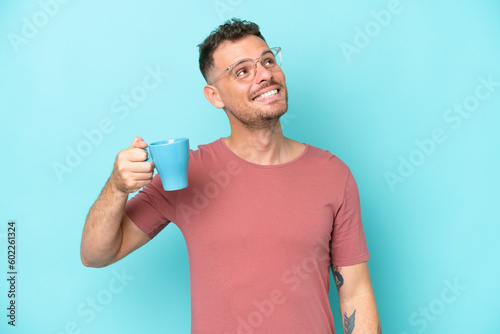 Young caucasian man holding cup of coffee isolated on blue background looking up while smiling