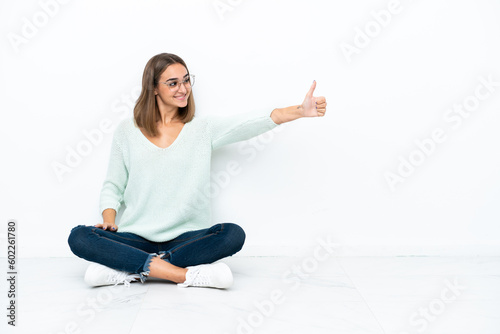 Young caucasian woman sitting on the floor isolated on white background giving a thumbs up gesture