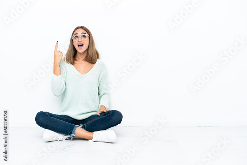 Young caucasian woman sitting on the floor isolated on white background pointing up and surprised
