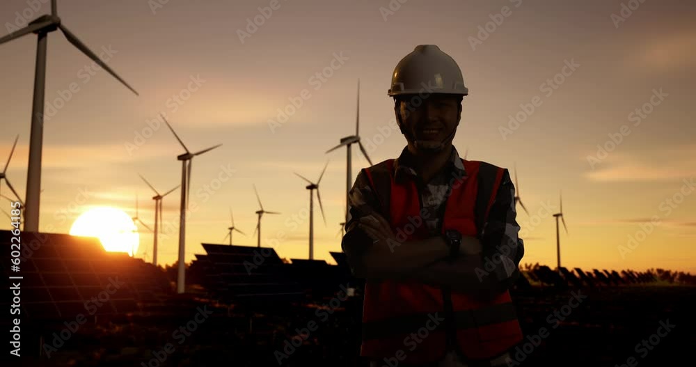 Asian Male Engineer In A Helmet Smiling And Posing With Arms Crossed While Silhouette In Front Of Wind Turbines Rotating At Sunset

