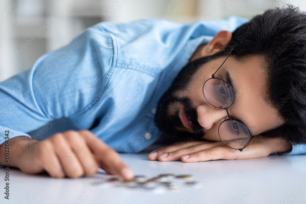 Financial Problems. Depressed Indian Man Counting Coins Lying On Table Financial Problems. Depressed Indian Man Counting Coins Lying On Table