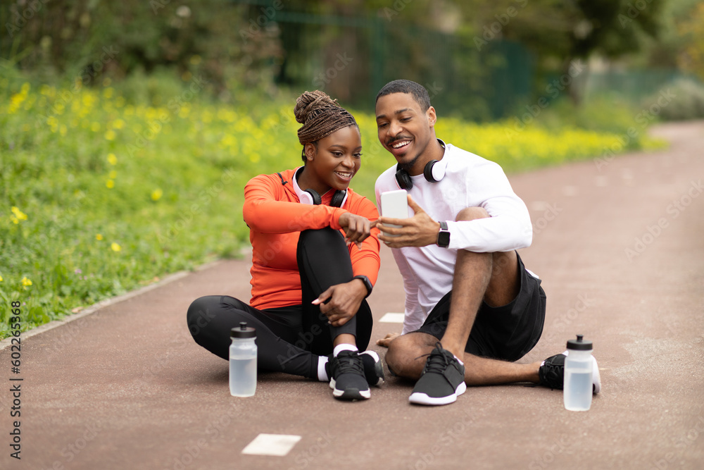 Happy young african american husband and wife sitting, talking with bottle of water, chatting in social networks - obrazy, fototapety, plakaty 