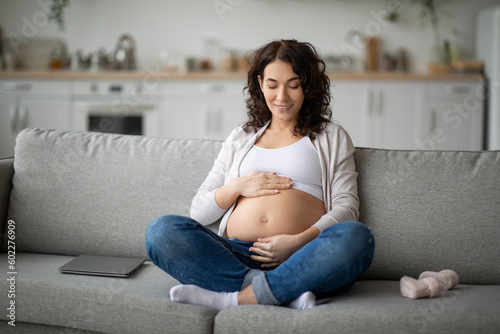 Smiling pregnant woman caressing her tummy while relaxing on couch at home