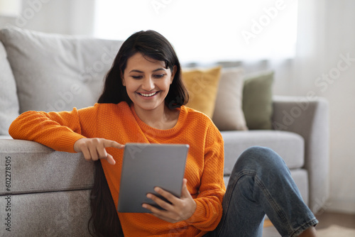Happy eastern woman sitting on floor at home, using tablet