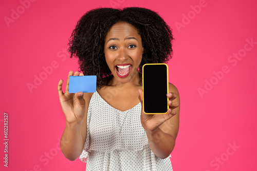 Happy surprised millennial african american curly lady show credit card and smartphone with blank space