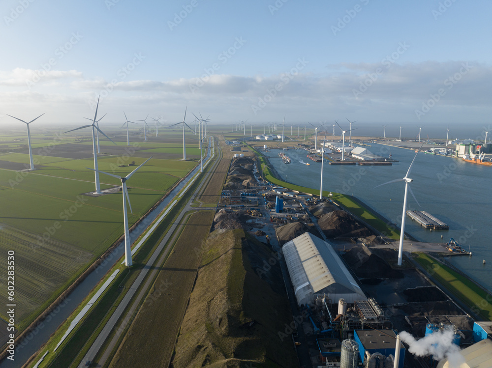 Fototapeta premium Discover the fascinating connection between wind energy and maritime trade with an unforgettable aerial footage of Eemshaven port, showcasing the harmonious coexistence of wind turbines, ships, and