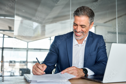Happy satisfied middle aged professional business man executive ceo manager, lawyer wearing suit sitting at desk signing law document writing signature making legal agreement corporate deal in office.