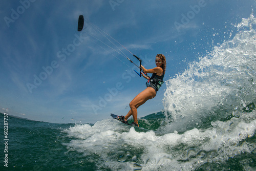 Kitesurfing girl in black sexy swimsuit with kite in sky on board in blue sea riding waves with water splash. Recreational activity, water sports, action, hobby and fun in summer time. Kiteboarding sp