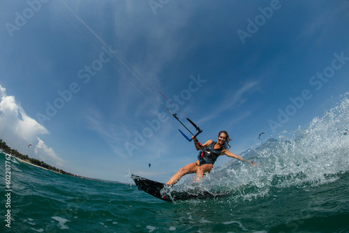 Kitesurfing girl in black sexy swimsuit with kite in sky on board in blue sea riding waves with water splash. Recreational activity, water sports, action, hobby and fun in summer time. Kiteboarding sp