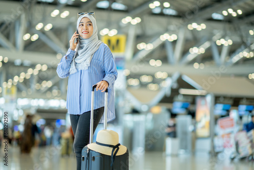 An Asian Muslim wearing a blue hijab is preparing for a vacation and she is at the airport. She is using her mobile phone to contact her friends and Muslim travelers.