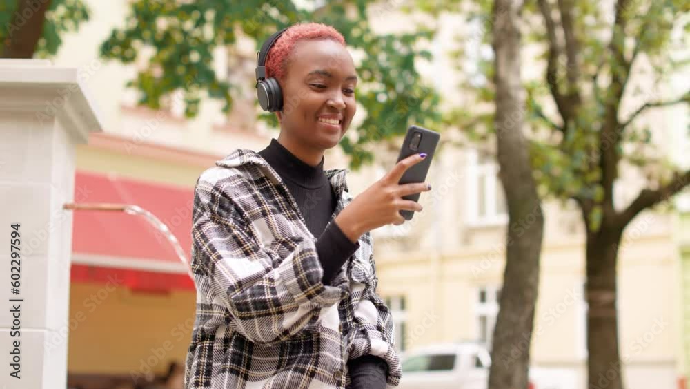 Portrait of happy young woman smiling looking at smartphone screen and listening to music in headphones. Pretty stylish female enjoying song in headphones using cellphone outdoor. Leisure concept Portrait of happy young woman smiling looking at smartphone screen and listening to music in headphones. Pretty stylish female enjoying song in headphones using cellphone outdoor. Leisure concept