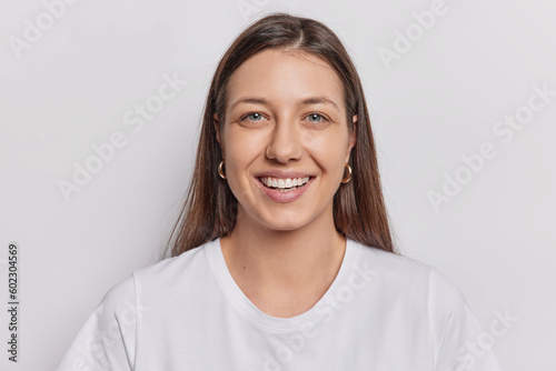 Φωτογραφία Portrait of good looking woman with dark straight hair smiles broadly expresses positive emotions wears casual t shirt isolated over white wall