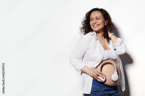 Young relaxed pregnant woman, future mom with tummy wearing white casual shirt, puts headphones on belly for baby to listen to music, smiling isolated on white background. Maternity pregnancy concept.
