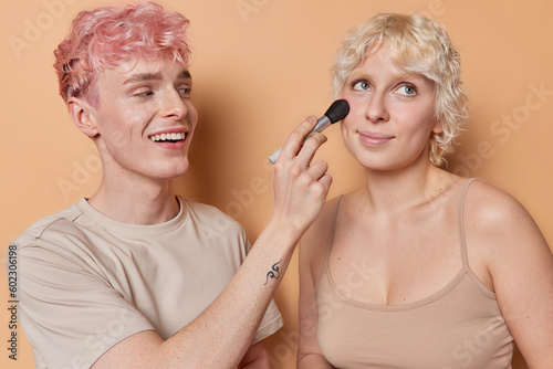 Happy freckled pink haired boyfriend uses professional cosmetic brush applies facial powder on girldfriends face puts on daily makeup have fun together learns some beauty procedures smiles gladfully.