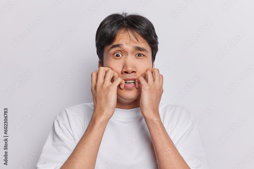 Scared anxious man stares with terror at camera keeps hands near mouth dressed in casual t shirt isolated over white background looks with panic. Human reactions and negative emotions concept Scared anxious man stares with terror at camera keeps hands near mouth dressed in casual t shirt isolated over white background looks with panic. Human reactions and negative emotions concept