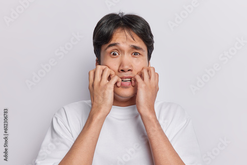 Scared anxious man stares with terror at camera keeps hands near mouth dressed in casual t shirt isolated over white background looks with panic. Human reactions and negative emotions concept © wayhome.studio 