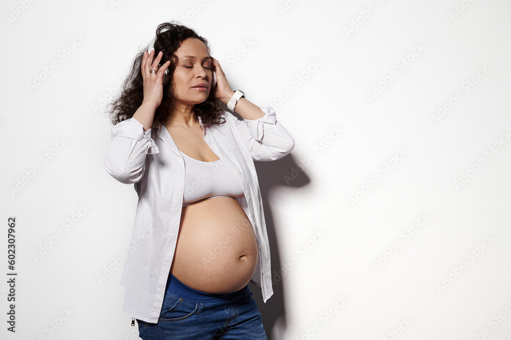 Photo studio portrait of delightful pregnant woman showing her belly, wearing wireless headphones, isolated on white background. People. Lifestyles. Pregnancy. Maternity. Leisure activity. Copy space Photo studio portrait of delightful pregnant woman showing her belly, wearing wireless headphones, isolated on white background. People. Lifestyles. Pregnancy. Maternity. Leisure activity. Copy space