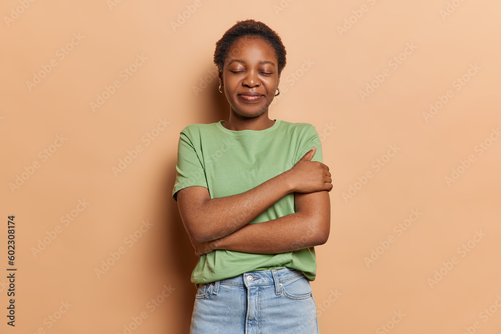 Indoor shot of dark skinned teenage girl embraces own body with arms keeps eyes closed dressed in casual green t shirt and jeans isolated over brown background recalls something. Love yourself concept Indoor shot of dark skinned teenage girl embraces own body with arms keeps eyes closed dressed in casual green t shirt and jeans isolated over brown background recalls something. Love yourself concept