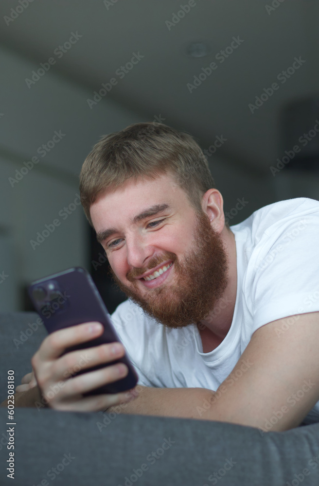 Young happy positive adult man relax, on sofa using smartphone for online leisure. People good well rest on lazy day at home or apartment concept. Young happy positive adult man relax, on sofa using smartphone for online leisure. People good well rest on lazy day at home or apartment concept.