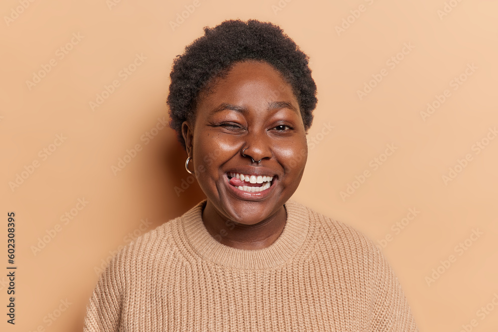 Young dark skinned cheerful woman winks eye and sticks out toungue has cheerful expression has playful expression wears casual knitted jumper isolated over beige background. Human facial expressions Young dark skinned cheerful woman winks eye and sticks out toungue has cheerful expression has playful expression wears casual knitted jumper isolated over beige background. Human facial expressions