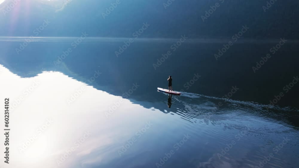 Epic and cinematic serene aerial shot of single paddle board move through still fjord water in Norway. Stand up paddle board in amazing scenery. Drone shot of paddle board in fjord