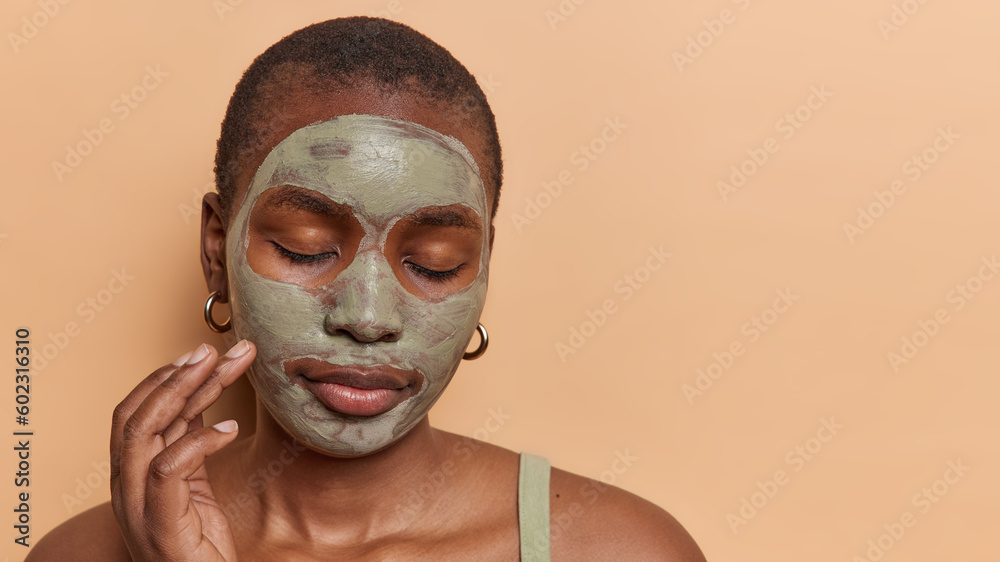 Headshot of dark skinned female model applies facial beauty mask to reduce pores keeps eyes closed enjoys skin care treatments stands bare shoulders isolated over brown background copy space for text - obrazy, fototapety, plakaty 
