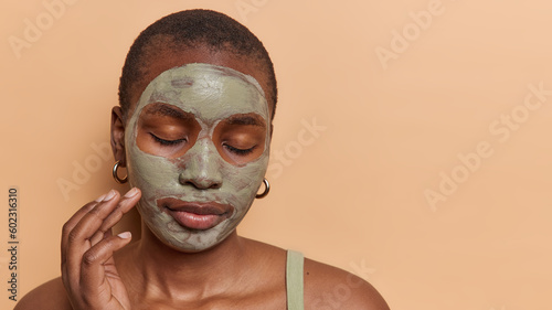 Headshot of dark skinned female model applies facial beauty mask to reduce pores keeps eyes closed enjoys skin care treatments stands bare shoulders isolated over brown background copy space for text