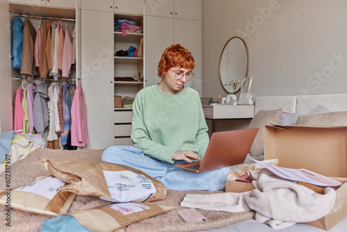 Serious red head woman uses laptop for selling clothes online makes order prepares parcels for customers dressed in casual clothes sits crossed legs on bed near wardrobe. People and resail fashion photo