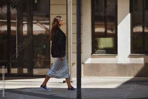 Beautiful brunette woman walks street city near buildings, wears luxurious attire clothes shiny maxi skirt with sequins, black sneakers and Tshirt. Light and shadow photo