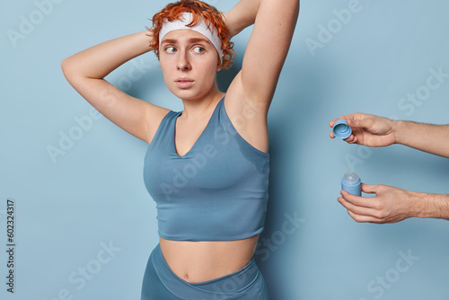 Embarrassed ginger sporty woman keeps arms raised up stands sweaty after hard training in gym being suggested new deodorant dressed in cropped top and leggings isolated over blue background.