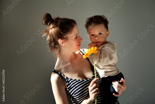 Portrait Beautiful Mother holds baby son and gerbera flower in hand while child looking at camera. Authentic portrait of real family, sunny, hard light on grey background, copy space © Anna Zhuk