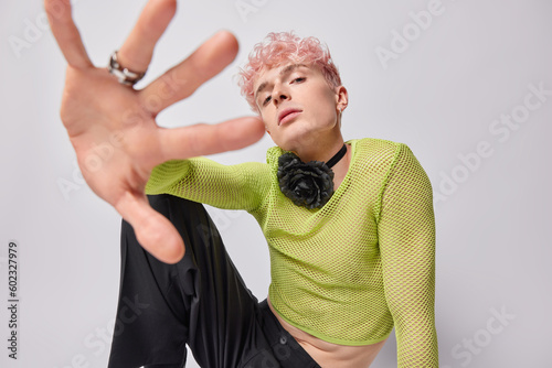 Photos Serious self confident androgynous pink haired man keeps hand towards camera wears green net jumper trousers poses against white background
