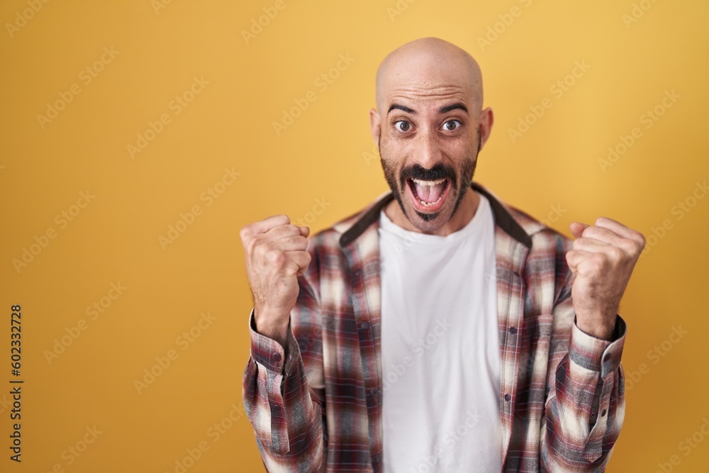 Naklejka premium Hispanic man with beard standing over yellow background celebrating surprised and amazed for success with arms raised and open eyes. winner concept.
