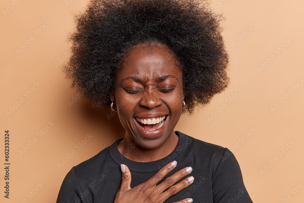 Φωτογραφία Overjoyed dark skinned woman keeps hand on chest laughs positively over something very funny keeps eyes cloosed dressed in black t shirt isolated over brown background
