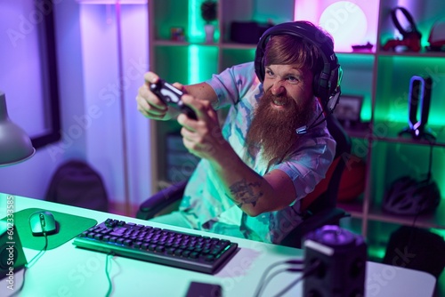 Young redhead man streamer playing video game using smartphone at gaming room