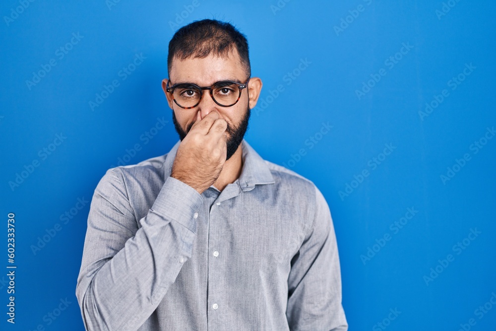 Middle east man with beard standing over blue background smelling something stinky and disgusting, intolerable smell, holding breath with fingers on nose. bad smell Middle east man with beard standing over blue background smelling something stinky and disgusting, intolerable smell, holding breath with fingers on nose. bad smell