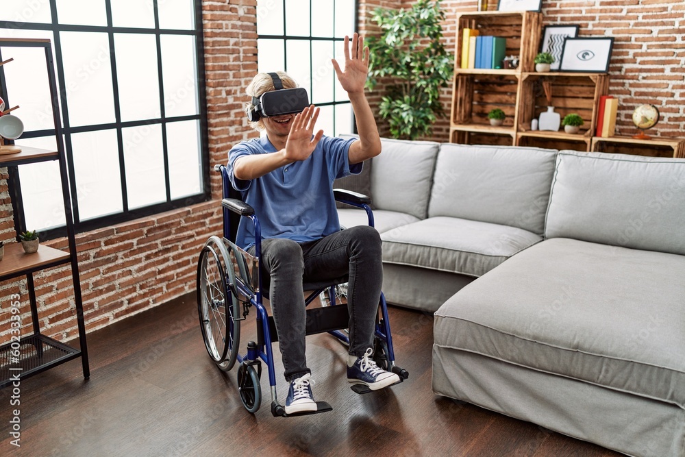 Young blond man playing video game using virtual reality glasses sitting on wheelchair at home