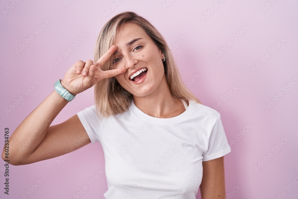 Young blonde woman standing over pink background doing peace symbol with fingers over face, smiling cheerful showing victory Young blonde woman standing over pink background doing peace symbol with fingers over face, smiling cheerful showing victory