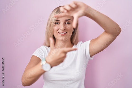 Young blonde woman standing over pink background smiling making frame with hands and fingers with happy face. creativity and photography concept.