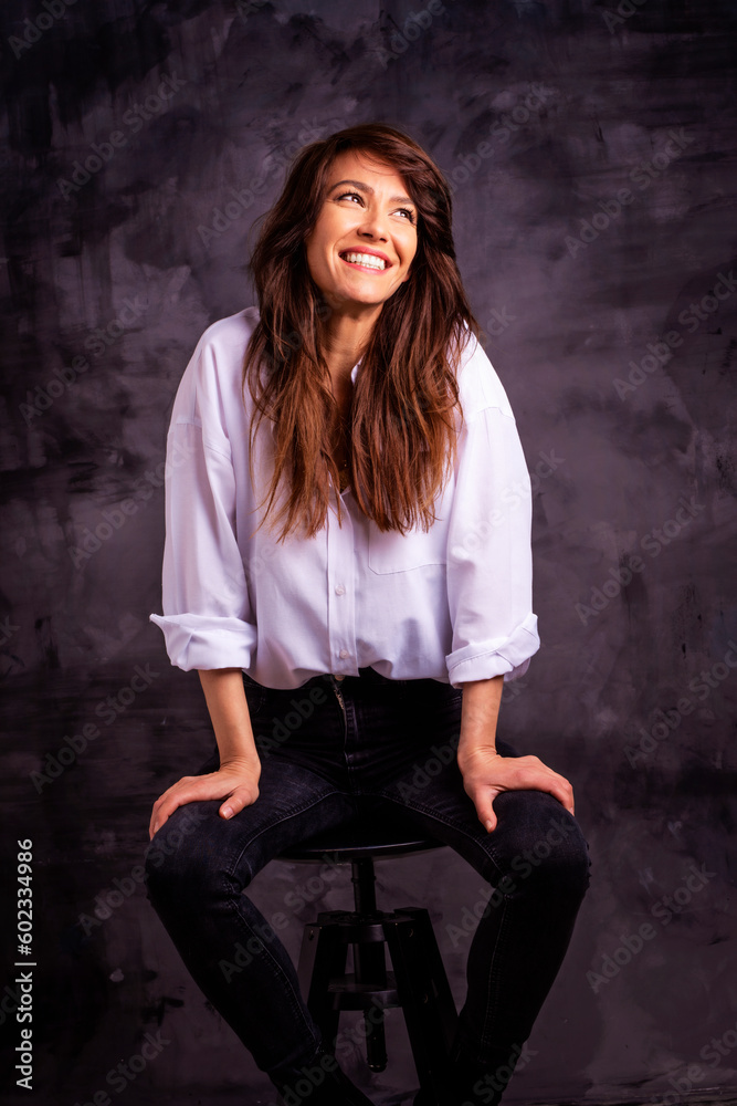 Fototapeta premium Studio portrait of a brunette haired attractive woman wearing white shirt and black jeans while posing at isolated dark background