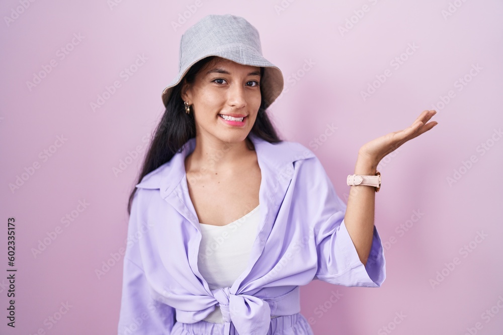 Young hispanic woman standing over pink background wearing hat smiling cheerful presenting and pointing with palm of hand looking at the camera. Young hispanic woman standing over pink background wearing hat smiling cheerful presenting and pointing with palm of hand looking at the camera.